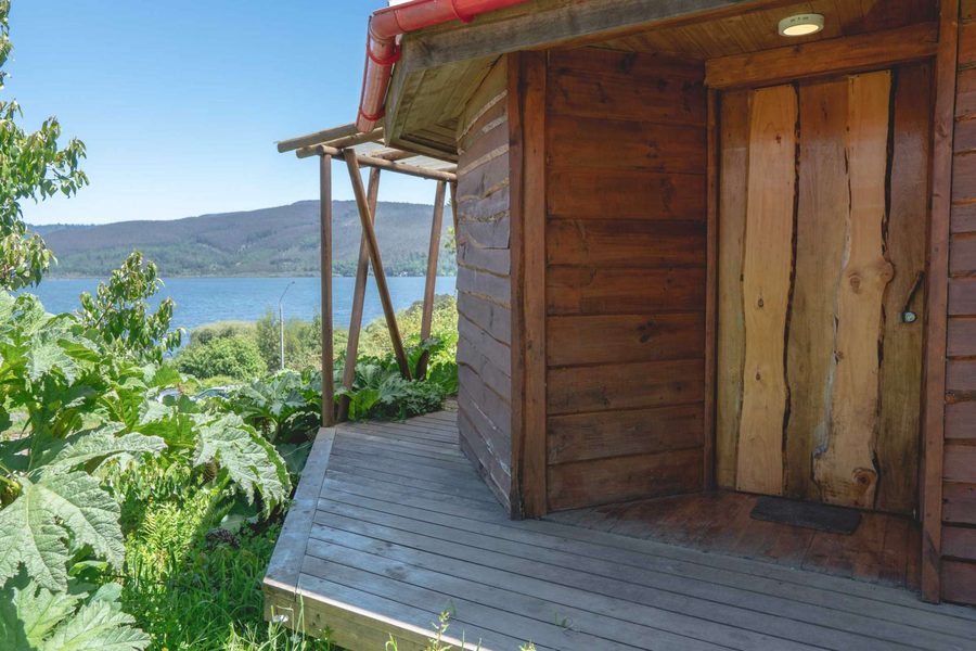 Cabaña de madera con terraza, rodeada de vegetación nativa y con el lago Lanalhue al fondo